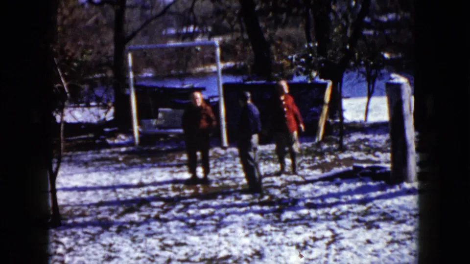 1960 home-movie frame shows three adults outdoors in snowy Wauconda, with the right figure raising an arm near the white goal frame.
