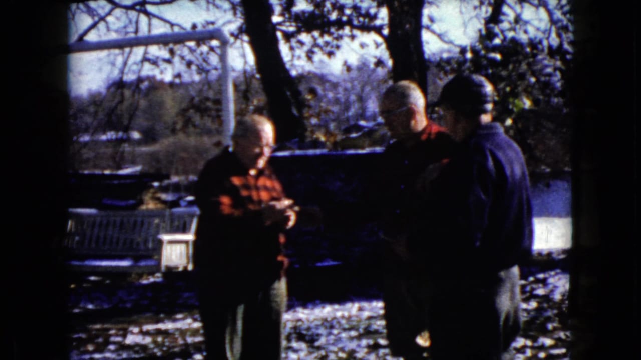 Three older men stand outdoors in snow, with one holding papers near a bench and water.