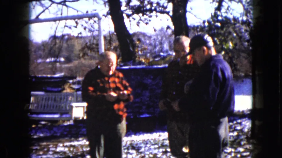 Three older men in winter clothing stand near snow and water in Wauconda, Illinois, with attention focused on papers or small items.