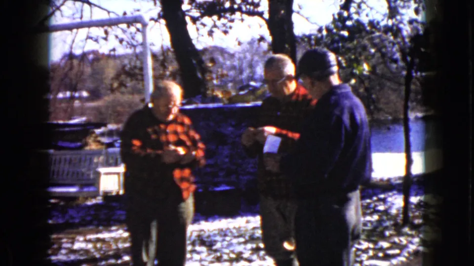 Older men in a snowy Wauconda outdoor setting appear to exchange or inspect papers, with bare trees and water behind them.