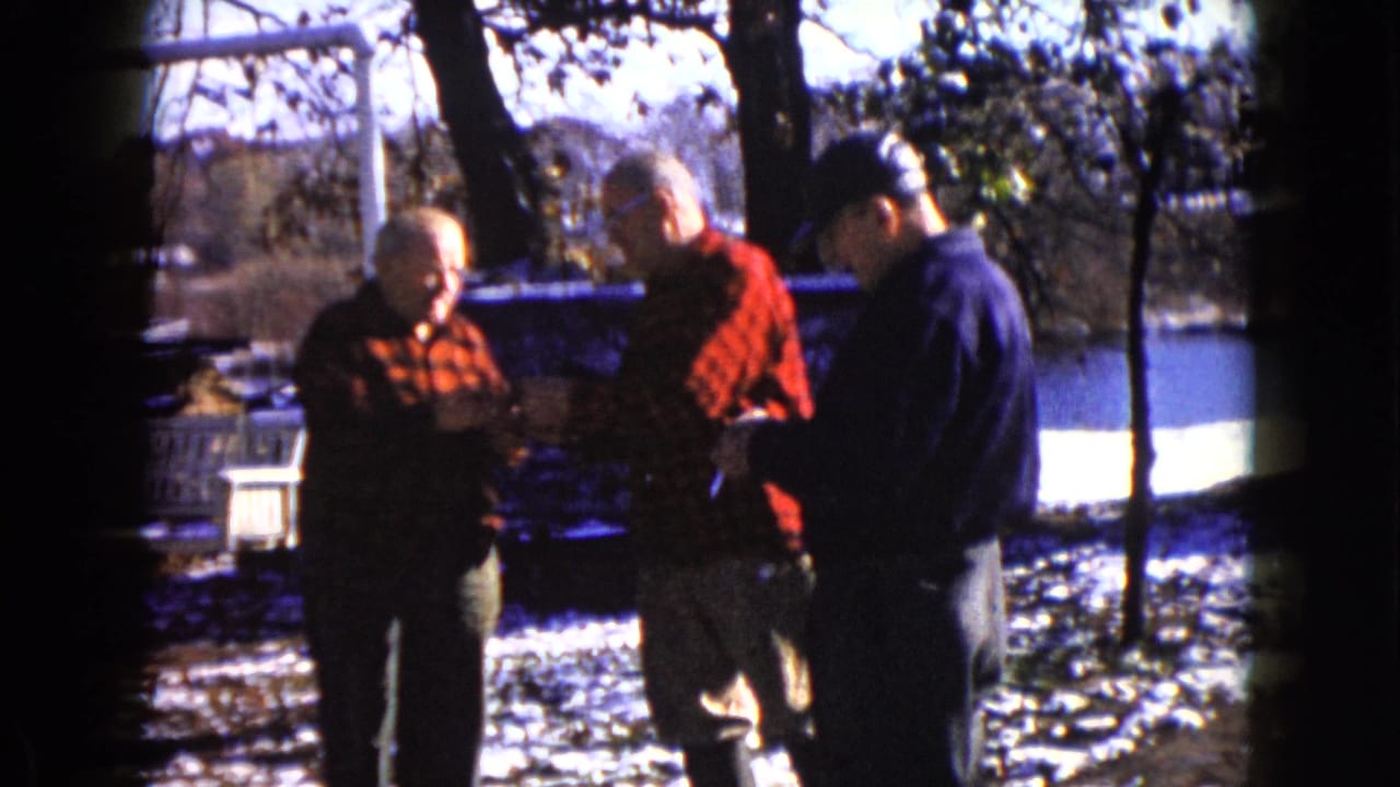 Three men stand outdoors near snow and bare trees, two wearing red plaid flannel shirts.
