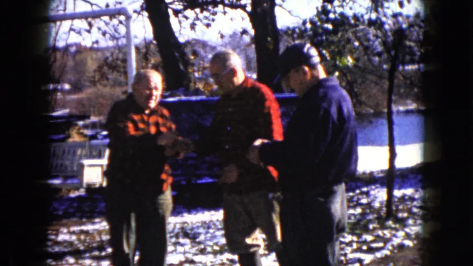 1960 Wauconda 8mm frame showing three men conversing in a winter yard, with flannel shirts, cap, snow, and trees behind them.