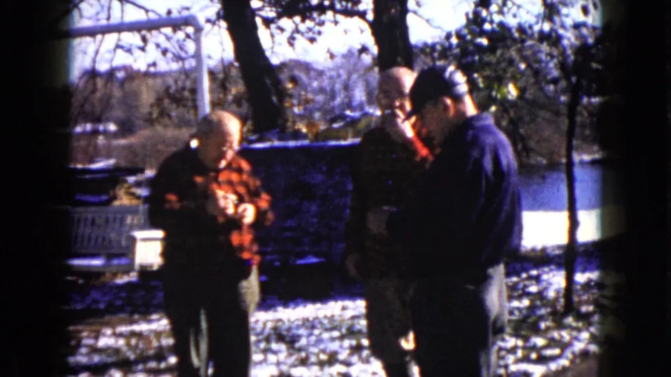 Home-movie frame from Wauconda, Illinois, showing three men in winter light with flannel shirts, snow patches, and bare trees.