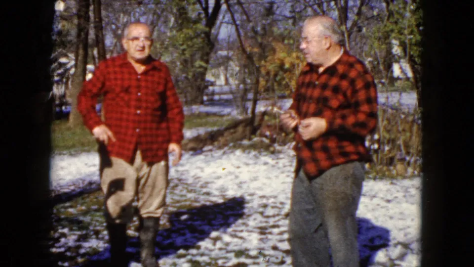 1960 Wauconda frame of two men in red plaid shirts standing in a snowy yard, with trees and winter ground visible behind them.