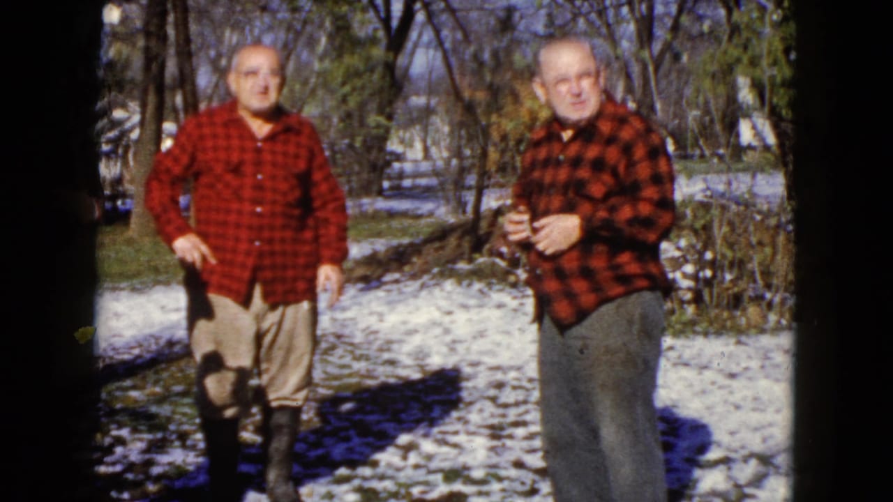 Two older men in red plaid shirts stand on snow-dusted ground near bare trees.