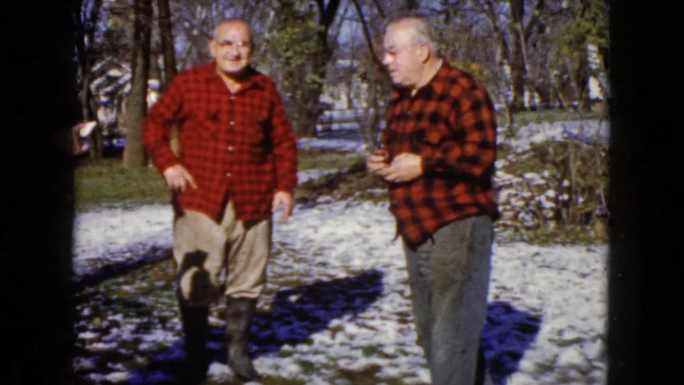 Two older men in matching red plaid shirts converse outdoors on snow-dusted ground in the stated 1960 Wauconda setting.