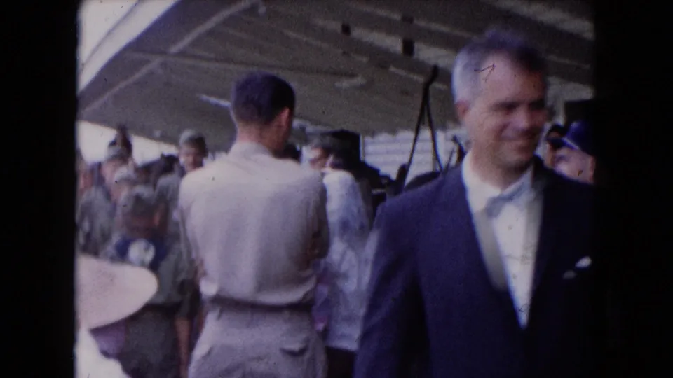 1961 8mm frame of a busy boat deck with a suited man, casually dressed passengers, and a canopy structure overhead.