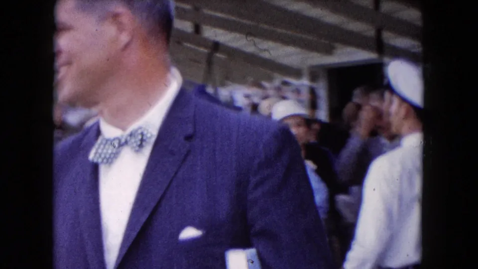 Buffalo 1961 home-movie frame showing a suited man with bow tie in profile amid crowded boat-deck passengers and uniform-like caps.