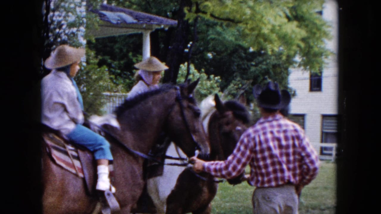 Two riders on horses beside a man in a plaid shirt and cowboy hat near houses and trees.