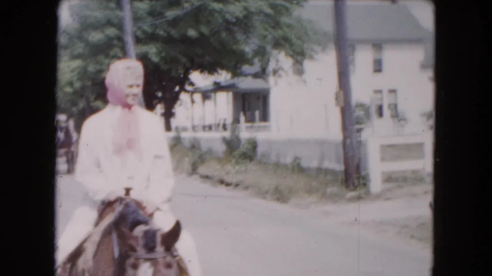 Woman in a pink kerchief rides a horse along a Buffalo residential street in the 1961 8mm footage.