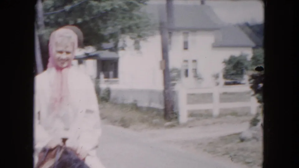 Closer 1961 Buffalo frame of the woman rider in a light outfit and pink kerchief passing a residential house and fence.