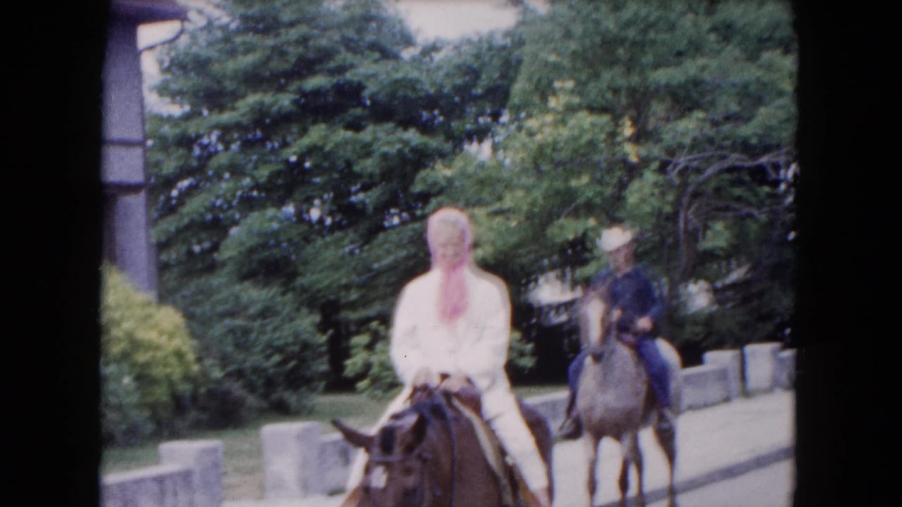 1961 Buffalo frame with a woman in white on a brown horse and a second rider in a hat behind her on a lighter horse.