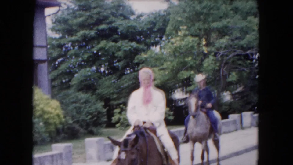 1961 Buffalo frame with a woman in white on a brown horse and a second rider in a hat behind her on a lighter horse.