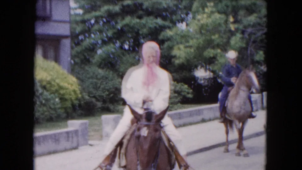 1961 Buffalo home-movie frame showing the foreground rider facing the camera on a brown horse, followed by a man on a lighter horse.
