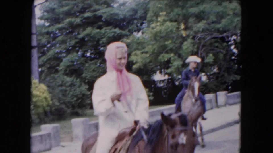 1961 Buffalo frame of a woman in white and pink headscarf passing close on a brown horse, with a second rider behind her.