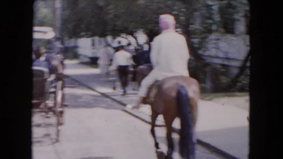 1961 Buffalo home-movie frame showing a rider on a brown horse moving along a tree-lined residential street behind people and a carriage.