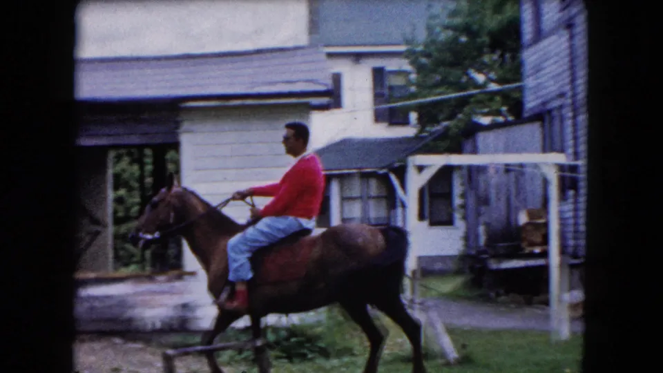 1961 Buffalo home-movie frame showing a man in a red sweater riding a brown horse through a residential yard area beside houses.