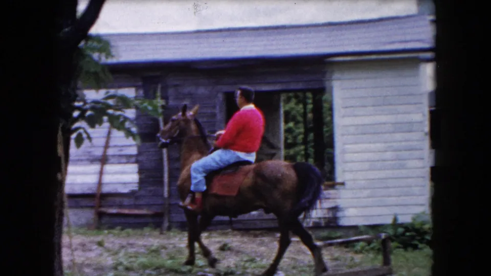 1961 Buffalo frame of a man in a red sweater riding a brown horse through a yard with light-colored residential buildings behind him.