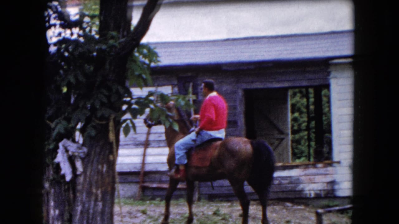 In 1961 Buffalo-area home-movie footage, a man in a red cardigan rides a brown horse past a tree and old wooden barn.
