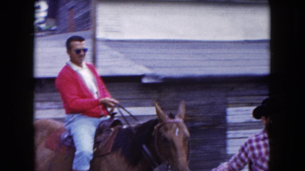 Man wearing sunglasses rides a brown horse past a weathered building.