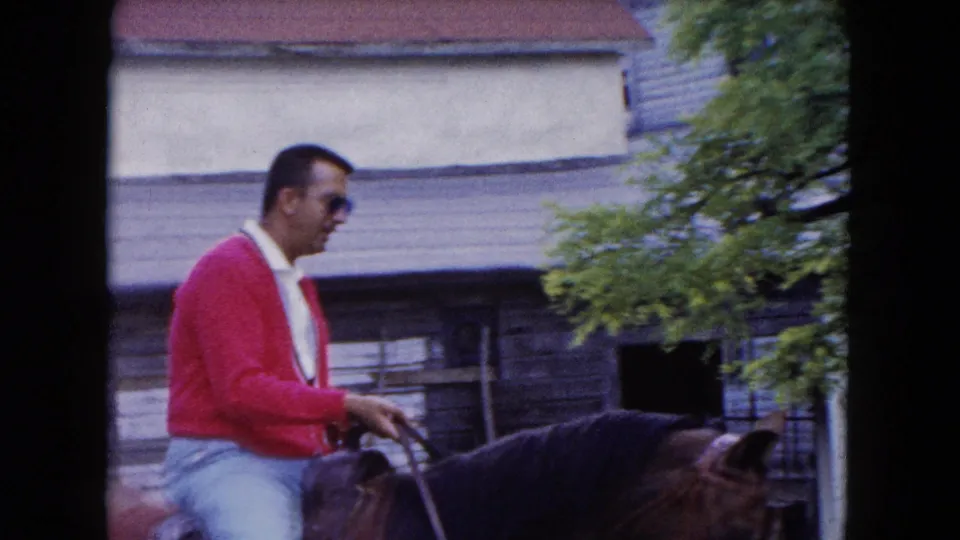 A closer side view shows the adult rider in a red cardigan guiding the horse beside weathered wooden siding in 1961 footage.