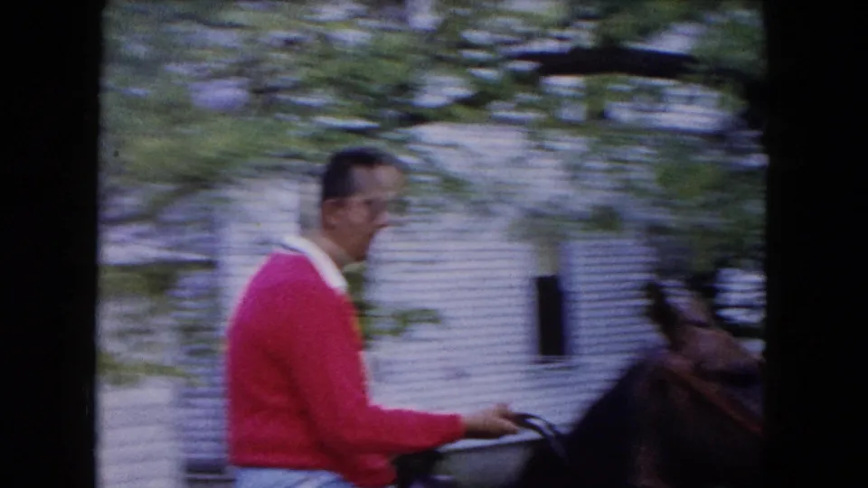 The rider moves under leafy branches on the brown horse, with a white house visible behind him in Buffalo, 1961.