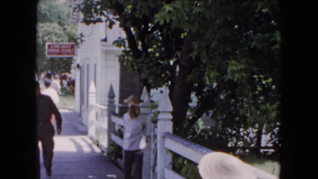 1961 Buffalo home-movie frame showing adults on a tree-shaded walkway beside a white building, railing, and Chicago Riding Stable sign.