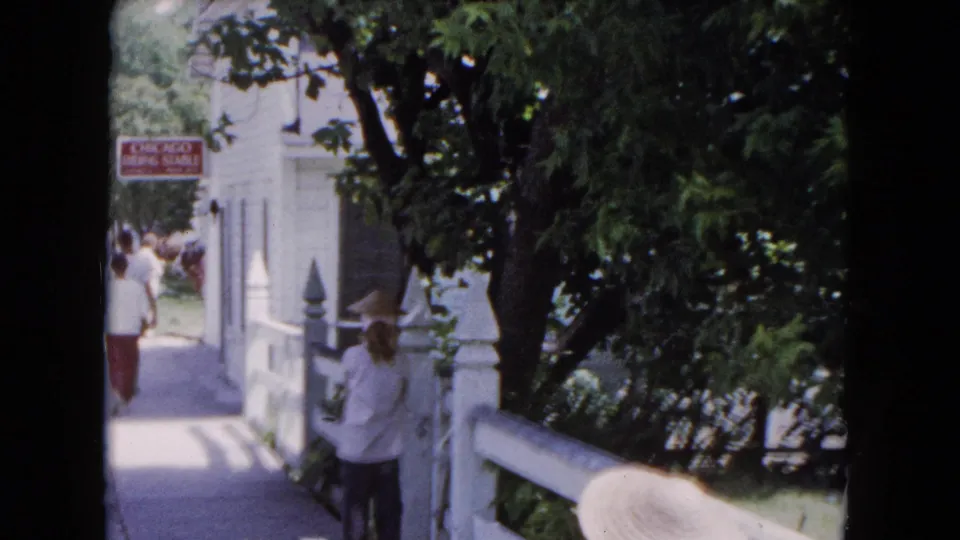 1961 Buffalo frame with a woman in a wide-brimmed hat near a white fence while adults move along a shaded walkway by the stable sign.