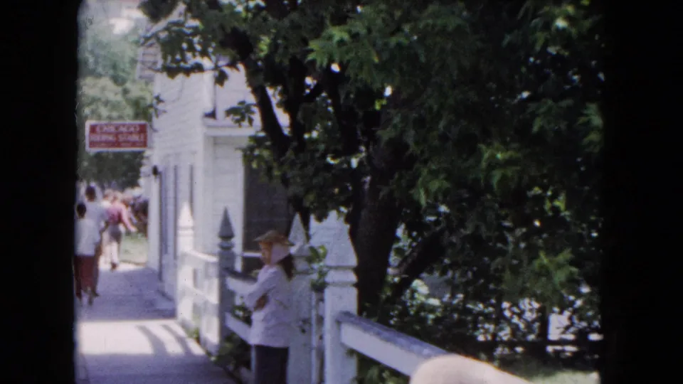 1961 Buffalo home-movie still showing adults walking near a white building and shaded trees, with the Chicago Riding Stable sign visible.