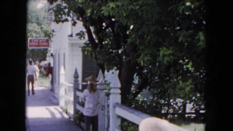 End frame from 1961 Buffalo footage with a woman near the white railing, leafy trees overhead, and adults walking toward the stable sign.