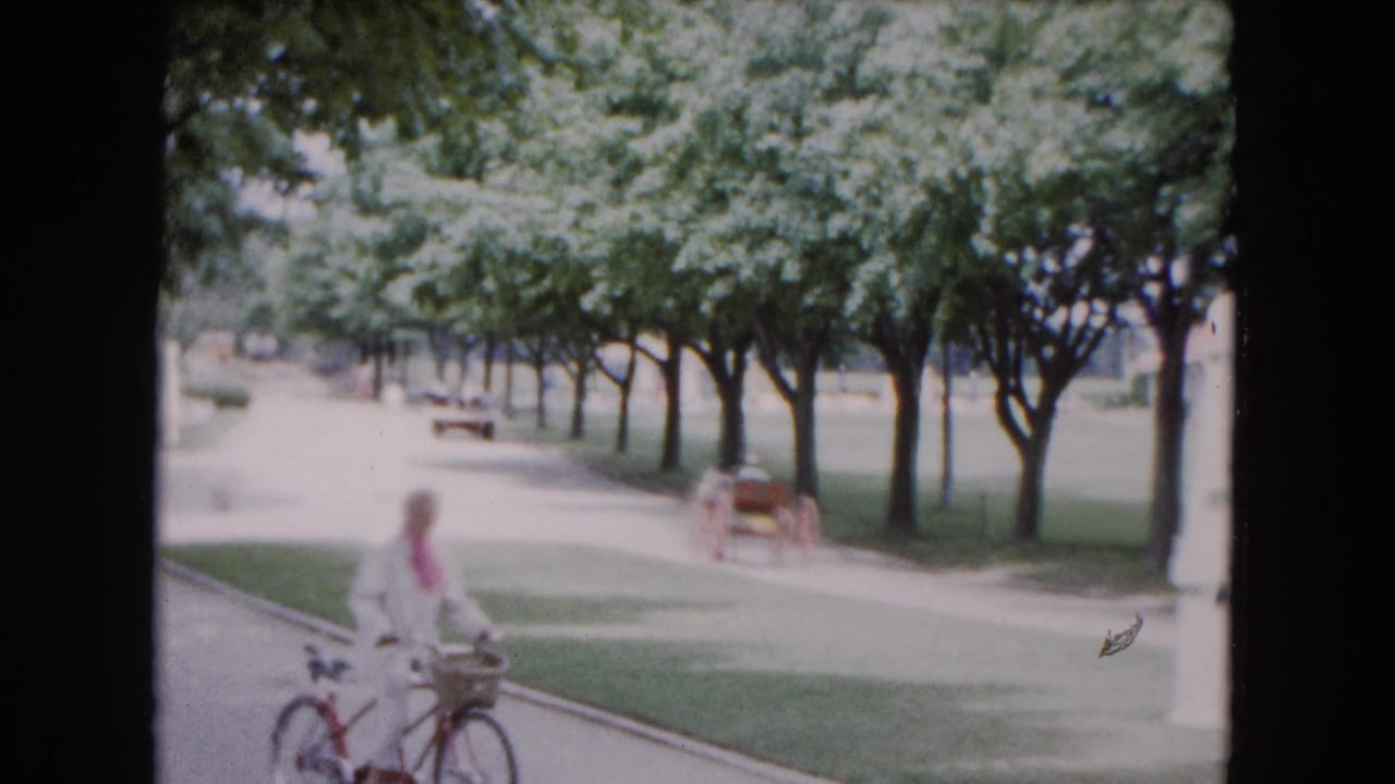 In 1961 Buffalo park footage, a woman in white with a pink scarf rides a bicycle on a paved path beneath trees.