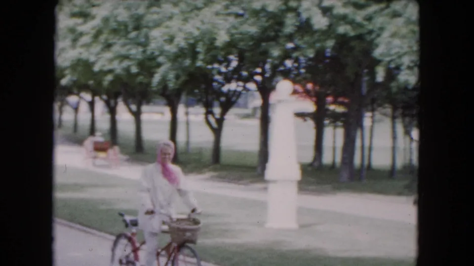 The woman faces the camera with her bicycle in a Buffalo park, with trees, lawn, and a white monument behind her.