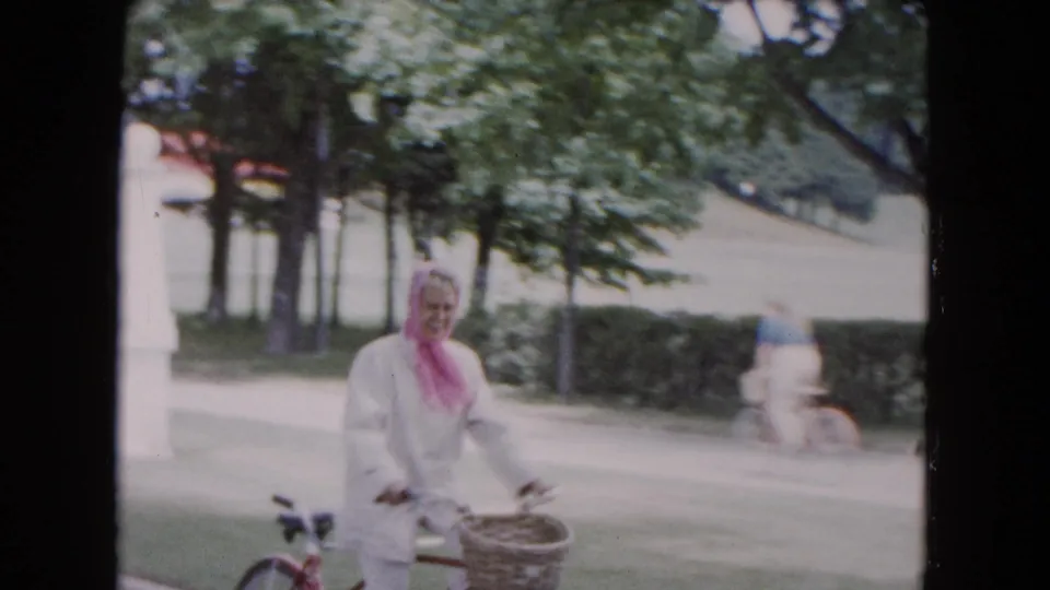 A woman in a white outfit and pink scarf smiles while riding a bicycle through the tree-lined Buffalo park setting.