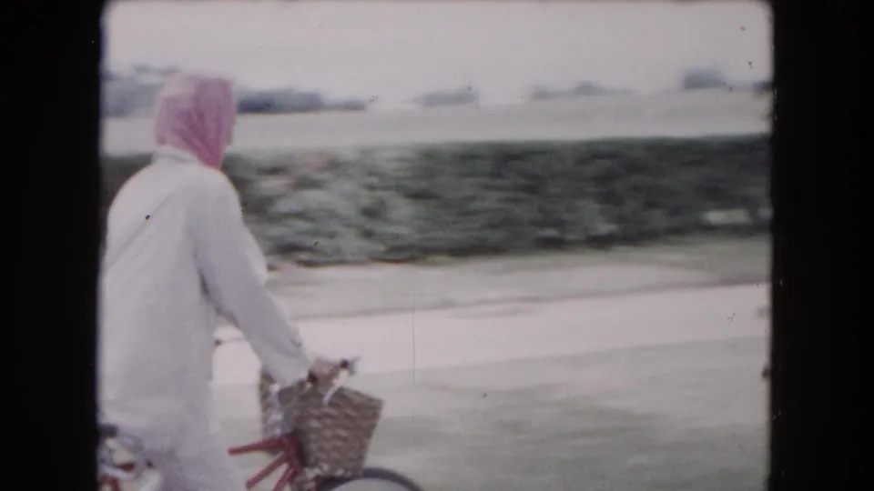 A side view shows the woman cycling with a front basket through the park, with lawn, hedge, and path visible in soft 8mm color.