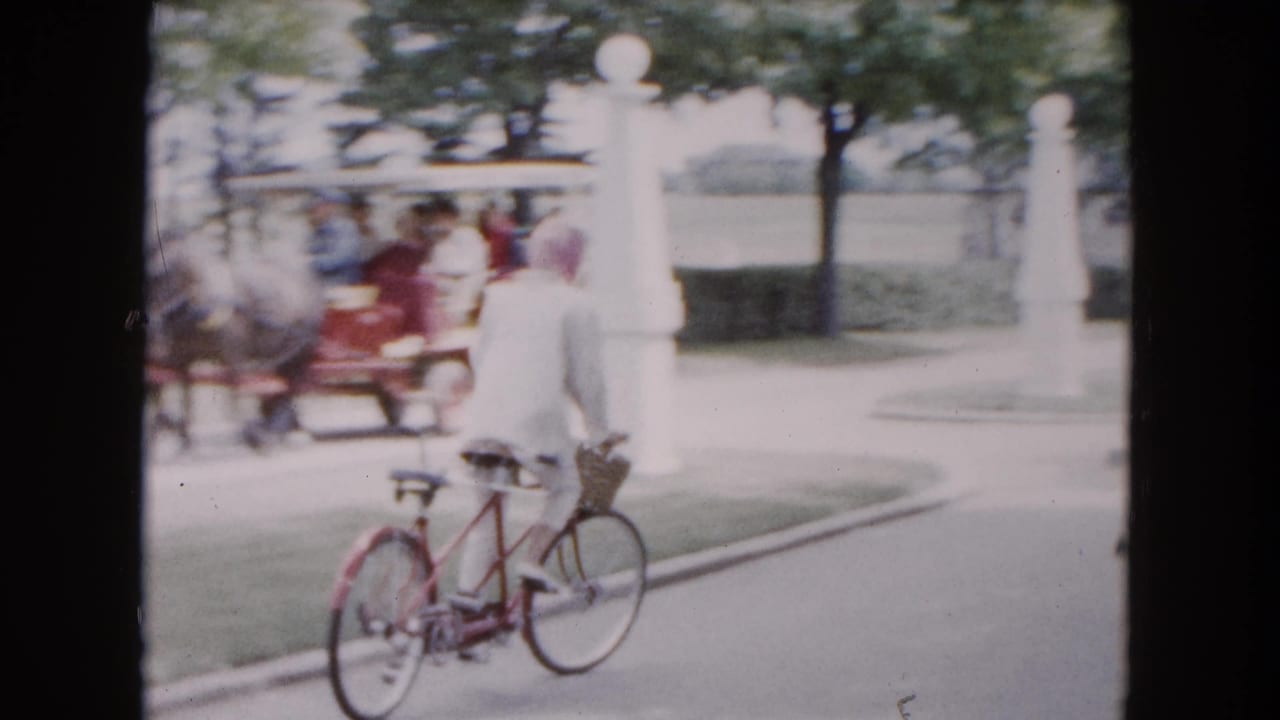The rider continues along a Buffalo road, with white entrance posts and a horse-drawn carriage visible beyond the bicycle.