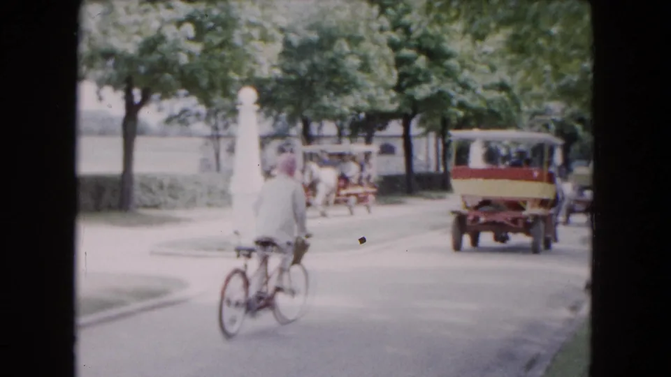 The 1961 Buffalo home-movie frame shows the cyclist from behind with a small vintage vehicle and carriage activity farther up the road.