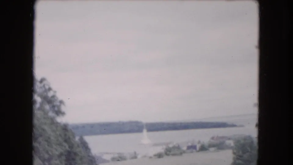 A soft 8mm frame shows a distant waterfront or lake view with a small sailboat and wooded shoreline, likely from the same Buffalo-area reel.