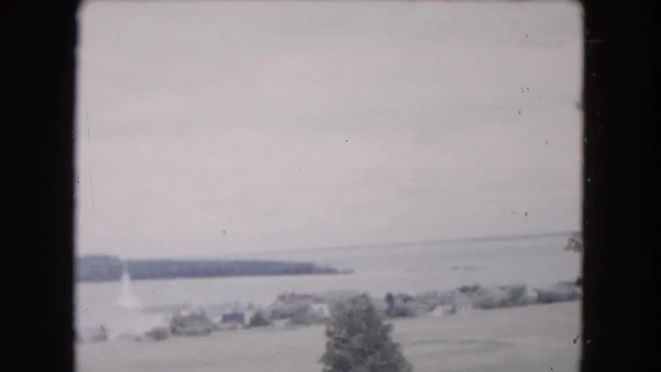 Faded 1961 8mm waterfront view near Buffalo, with a low wooded shoreline, open water, and blurred foreground trees.