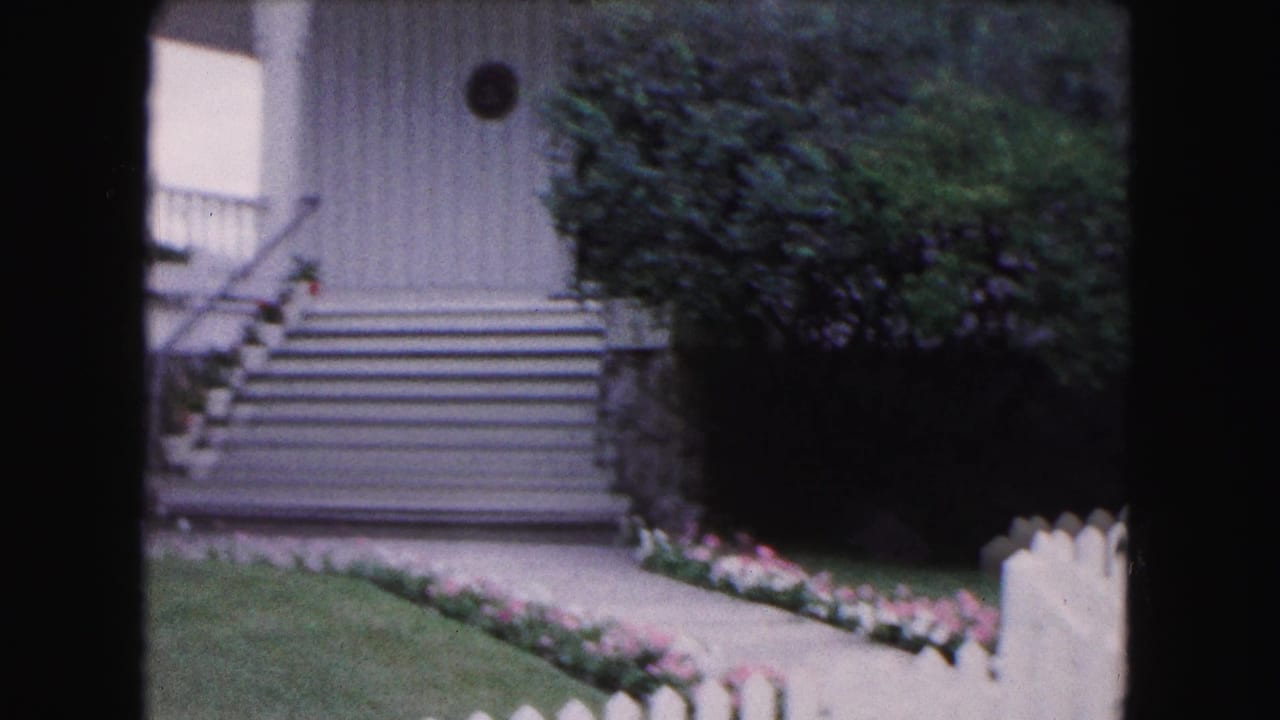 1961 Buffalo-area 8mm frame of stone steps, a pale house entrance, dense hedges, and flower borders behind a white fence.