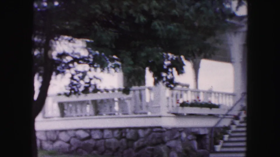 1961 Buffalo-area 8mm frame showing a porch railing, planter, stone foundation wall, stairs, and tree cover on the residence exterior.