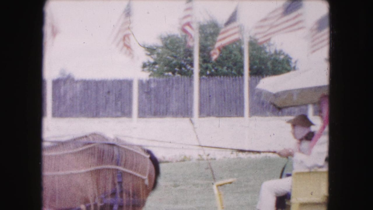 In 1961 Buffalo home-movie footage, a horse-drawn yellow carriage moves past American flags and a fenced outdoor area.