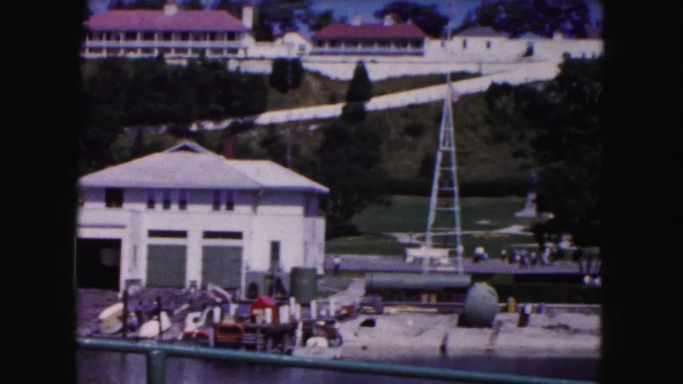 1961 Buffalo frame with a white boathouse-like structure, dock area, grassy park, tall white tower, and small distant pedestrians.