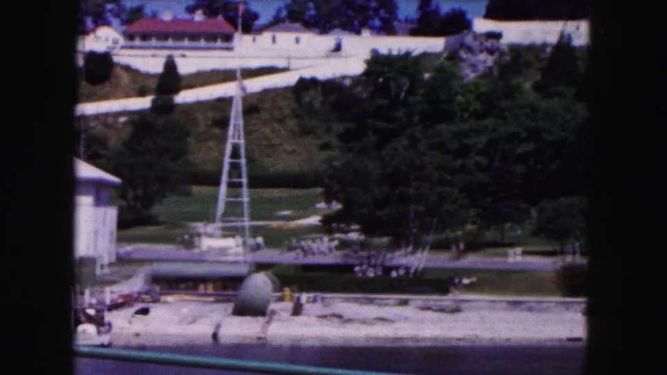 Buffalo home-movie frame showing a riverside park, white tower, trees, dock edge, green railing, and small figures walking in the distance.