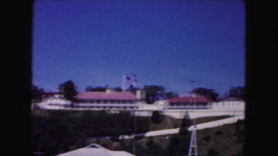 1961 Buffalo 8mm view of a red-roofed building with flagpoles, hillside landscaping, trees, paths, and a lattice tower.