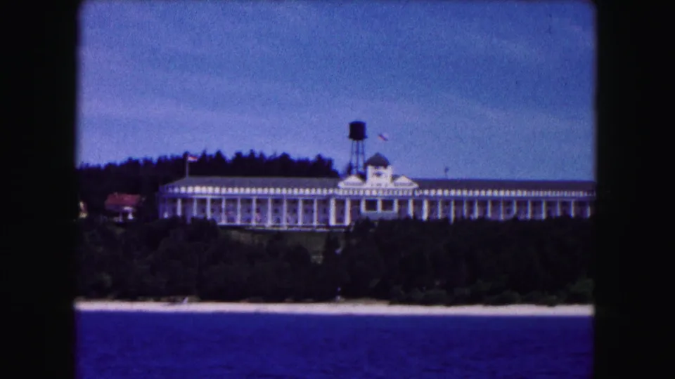 1961 Buffalo home-movie frame showing a white waterfront hotel-like building and water tower beyond trees and lake water.