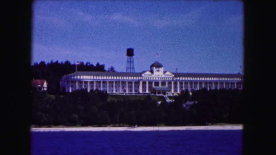 1961 Buffalo-area home-movie frame showing the white waterfront building and water tower from across blue water, with dark trees in front.