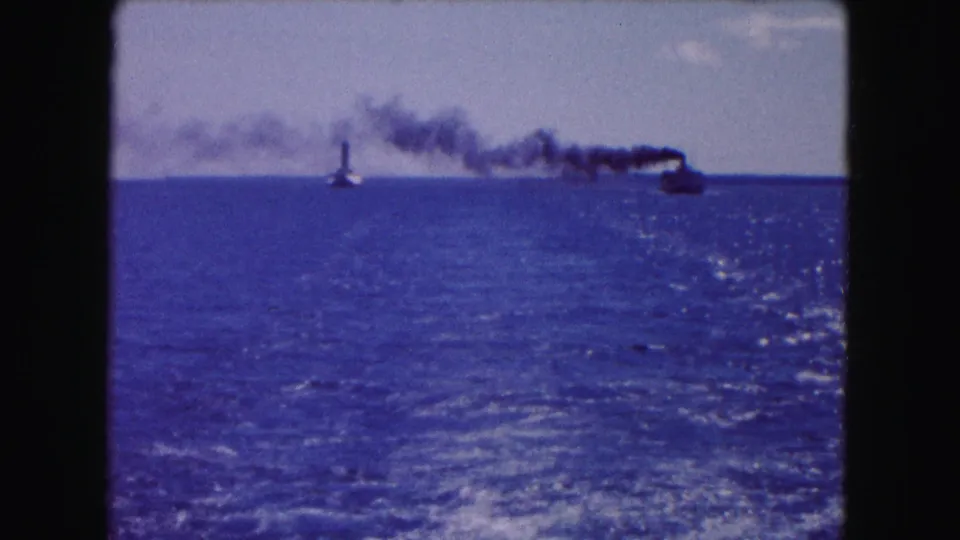 1961 Buffalo-area 8mm frame from a moving boat, with wake in foreground and smoke-trailing traffic in the distance.