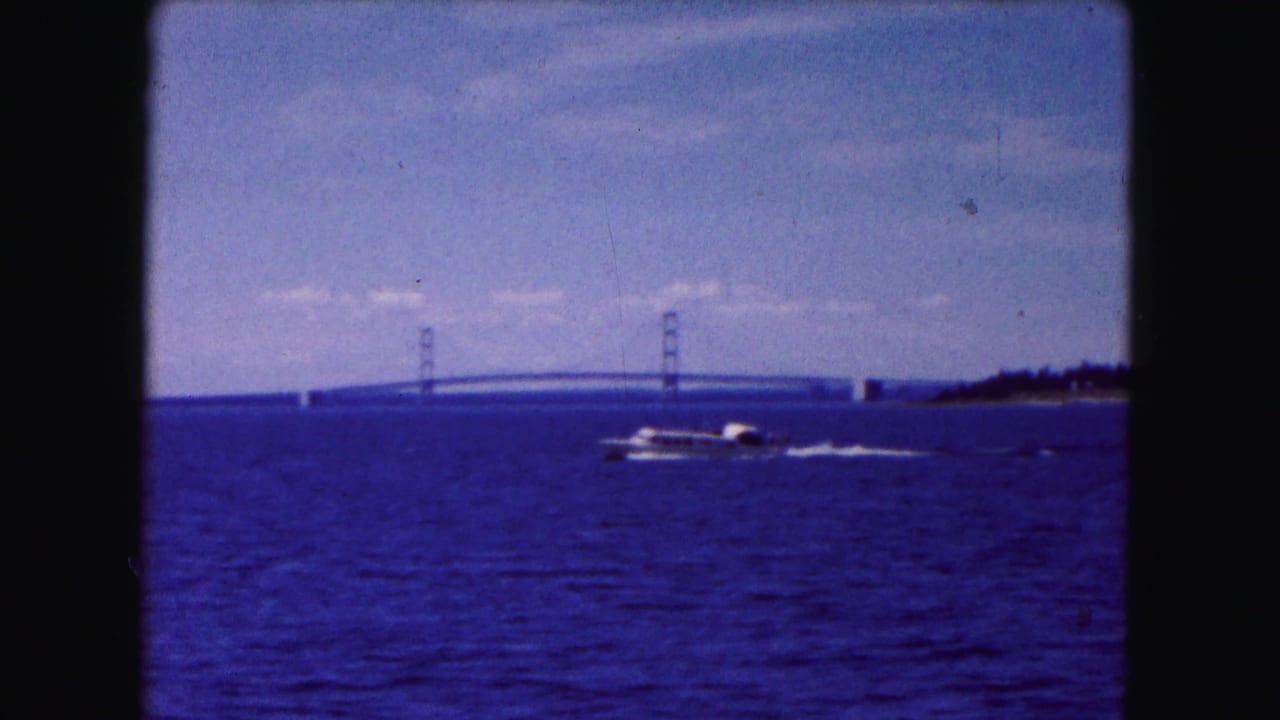 Motorboat crosses blue water with a suspension bridge and shoreline in the distance.