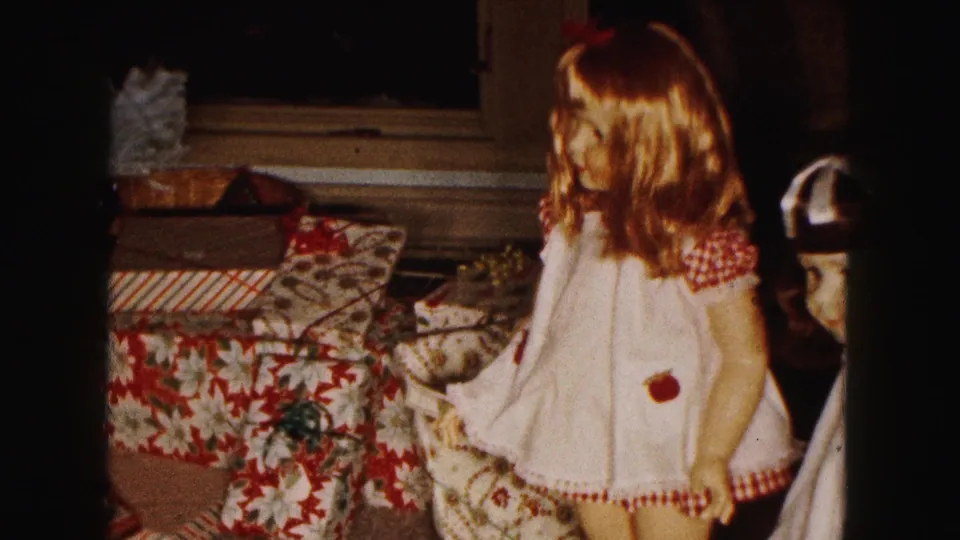 1961 Buffalo home-movie frame showing a brown-haired doll in a white dress near floral-wrapped Christmas gifts.