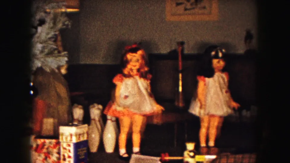 1961 Buffalo home-movie frame showing a child near the edge of a holiday setup with dolls, toy pins, gifts, and tree branches.